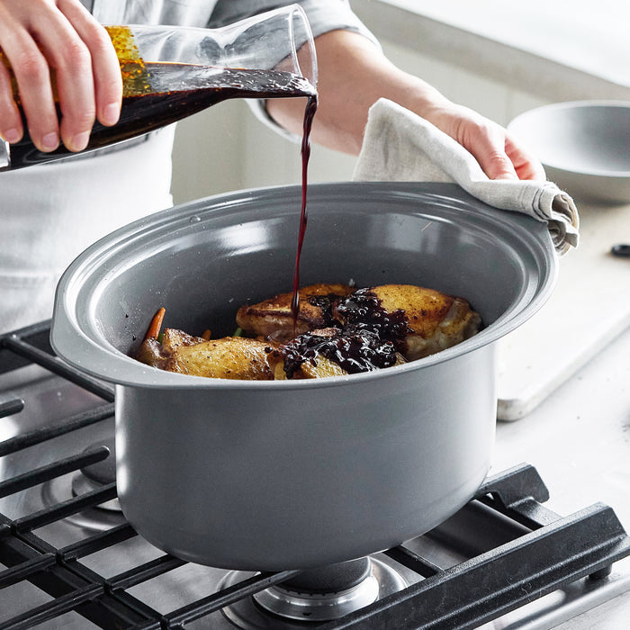 Red wine being poured over chicken in a clay 8-quart slow cooker pot on a stovetop.
