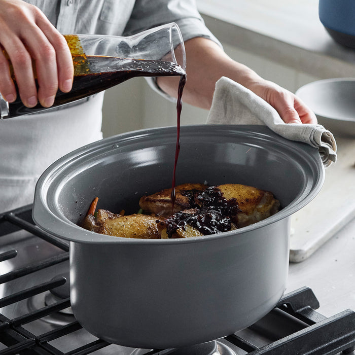 Red wine being poured over browned chicken in a silver slow cooker; cream-colored cookware on the stovetop nearby.