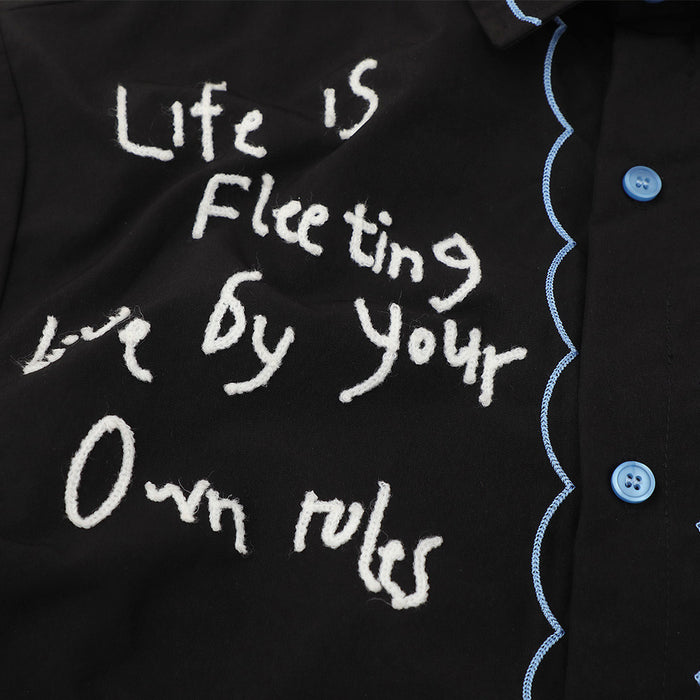 Black short sleeve shirt with white embroidered slogan, blue scalloped trim, and blue buttons