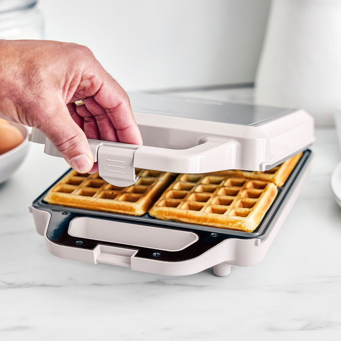 Person lifting a waffle from a white waffle maker on a light surface.