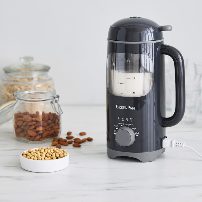 Graphite milk maker beside glass jars of nuts and a bowl of soybeans on a white kitchen counter.