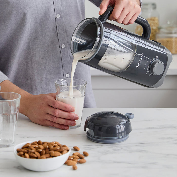 Milk is poured from a graphite Better Blend Milk Maker into a glass, with a bowl of almonds next to them.