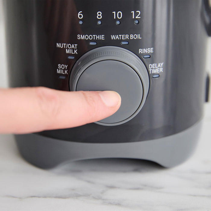 Hand pressing the large dial on a graphite Better Blend Milk Maker, dial set to smoothie, water boil, and soy milk options.