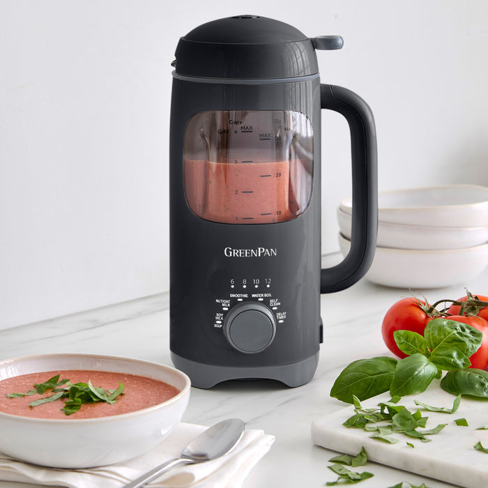 Graphite milk maker with tomato soup inside, next to a bowl of soup, fresh tomatoes, and spinach on a white counter.