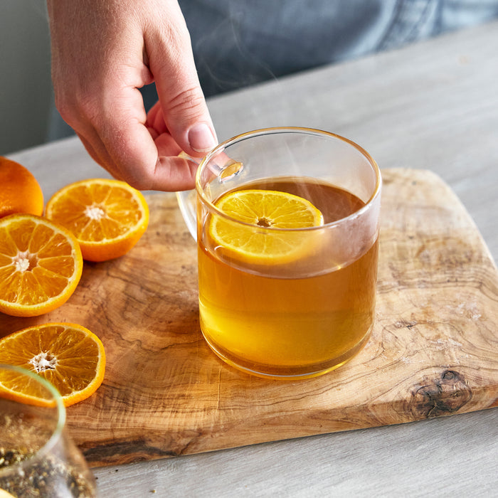 Hand holding a glass mug of tea with orange beside sliced oranges and a Better Blend Milk Maker on a wooden board.