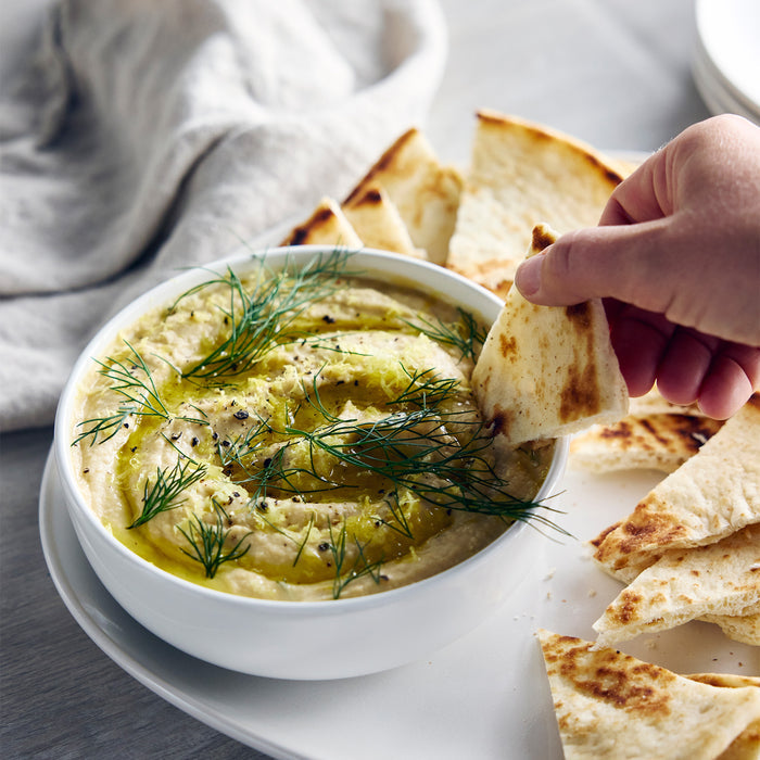 Pita being dipped into hummus with dill and olive oil, next to the Better Blend Milk Maker in graphite color.
