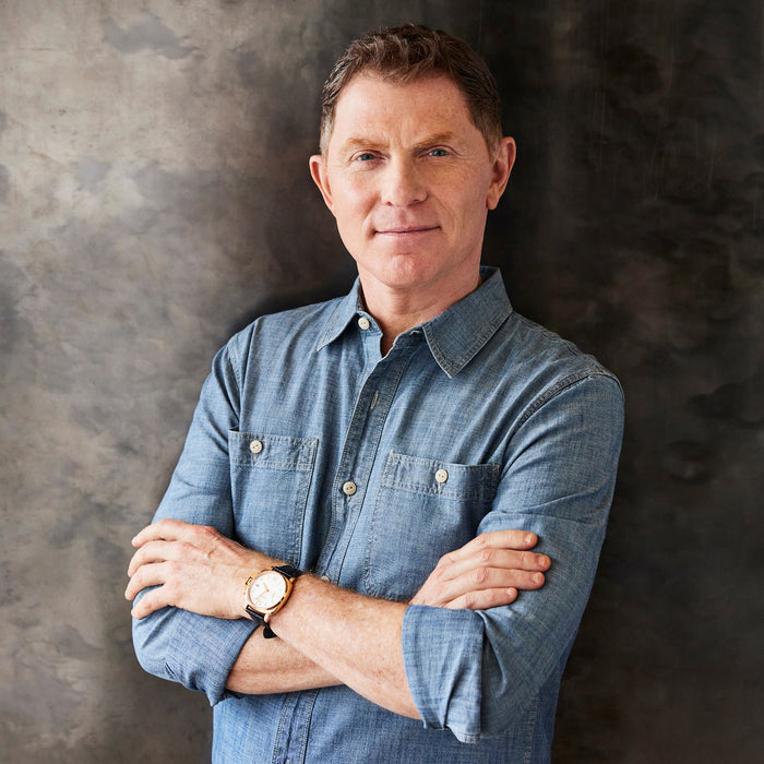 A man with short brown hair, wearing a blue button-up shirt and a watch, stands smiling with arms crossed before a textured dark gray background, ready to showcase the Bobby Flay by GreenPan 11" Carbon Steel Crepe Pan.