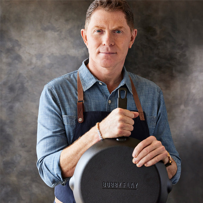 A man in a blue shirt and apron stands against a textured background, smiling as he holds the Bobby Flay by GreenPan 5-Piece Cast Iron Set with the Bobby at Home Cookbook, facing the camera.