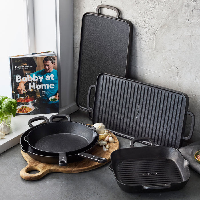 The Bobby Flay by GreenPan 4-Piece Cast Iron Set with Bobby at Home Cookbook, featuring a grill pan, griddle, skillet, and baking dish, is displayed on a gray kitchen counter with herbs and garlic.