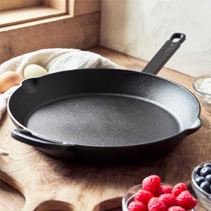 The Bobby Flay by GreenPan 5-Piece Cast Iron Set with Bobby at Home Cookbook sits on a wooden countertop near a window, with natural light illuminating eggs, flour, blueberries, and raspberries in glass bowls around it.