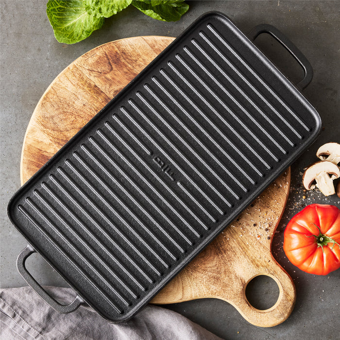 The Bobby Flay by GreenPan 5-Piece Cast Iron Set with Bobby at Home Cookbook is displayed on a round wooden board, surrounded by fresh veggies and a gray cloth atop a gray countertop.