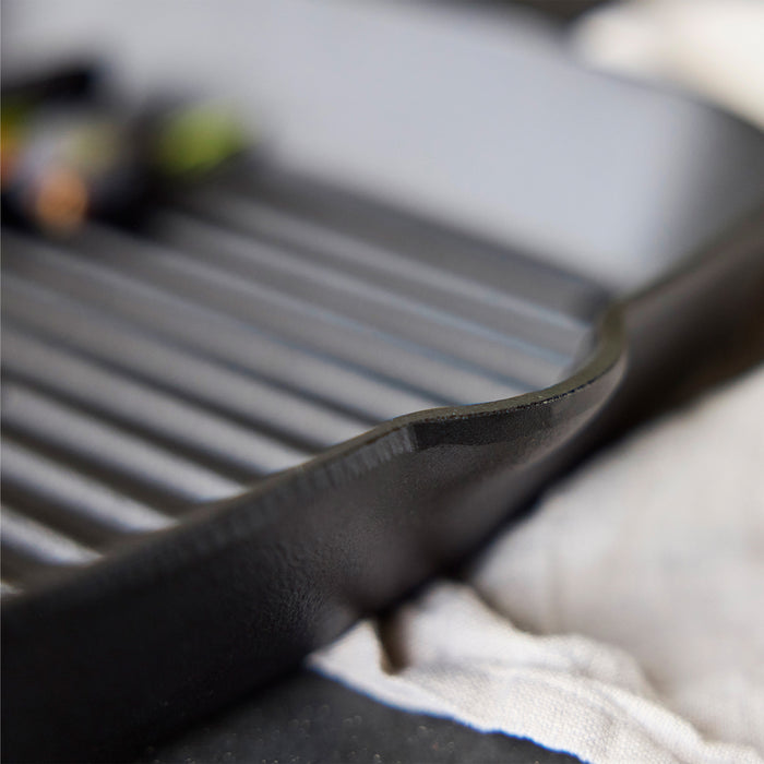 Close-up of the Bobby Flay by GreenPan 5-Piece Cast Iron Set, highlighting the spacious grill pan with ridges, textured edge, and heirloom-quality design—includes Bobby at Home Cookbook.
