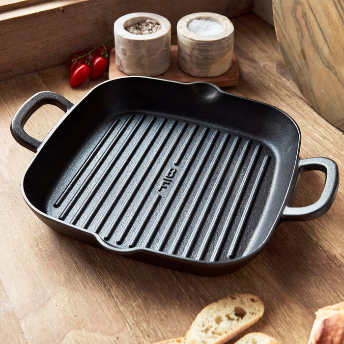 The Bobby Flay by GreenPan 5-Piece Cast Iron Set with Bobby at Home Cookbook, featuring a black square grill pan with two handles, rests on a wooden countertop near a window amid salt and pepper shakers, cherry tomatoes, and bread slices.