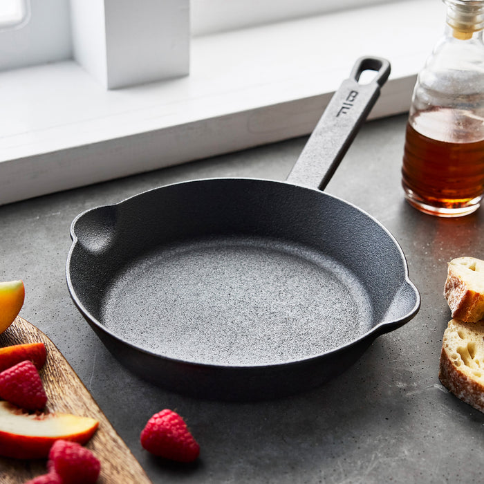 The Bobby Flay by GreenPan 6-Piece Cast Iron Set with Bobby at Home Cookbook sits on a gray countertop near a window, surrounded by syrup, bread, raspberries, and peach slices—perfect for daily use.