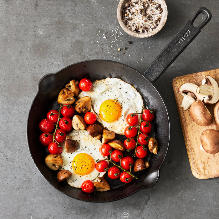 The Bobby Flay by GreenPan 8" and 10" Cast Iron Frypan Set features a cast iron skillet with sunny-side-up eggs, sautéed cherry tomatoes, and mushrooms on a gray surface. A wooden board beside it holds raw mushrooms plus a bowl of salt and pepper.