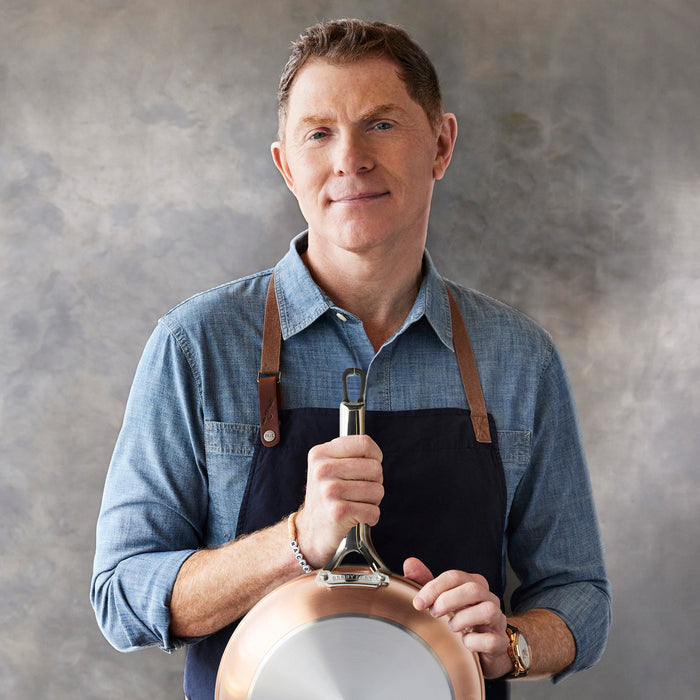 Man in blue shirt and apron holding a copper-clad Bobby Flay by GreenPan pan against a gray background.
