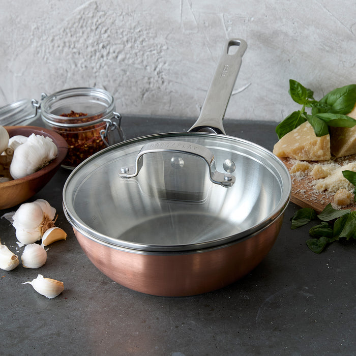 Copper-clad 2-quart chef’s pan with lid on a kitchen counter, next to garlic and spice jars.