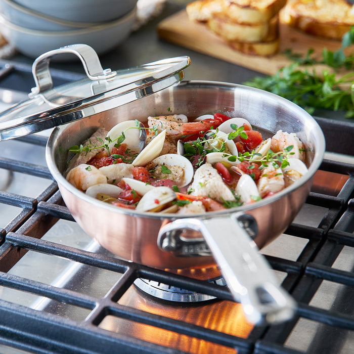Copper-clad 2-quart chef’s pan with lid slightly open, simmering seafood, tomatoes, and herbs.