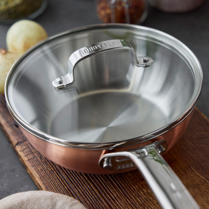 Copper chef’s pan with lid on a wooden board, surrounded by onions and assorted spices.
