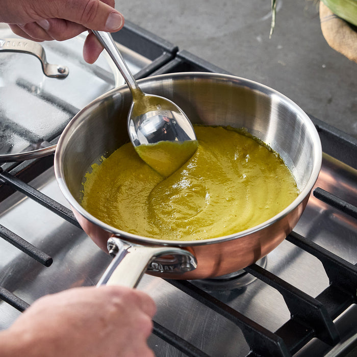 Green sauce being stirred with a metal spoon in a copper-clad chef’s pan on a stovetop.