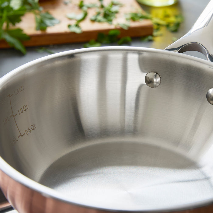 Copper-clad 2-quart chef’s pan with lid, blurred herbs and cutting board in the background.
