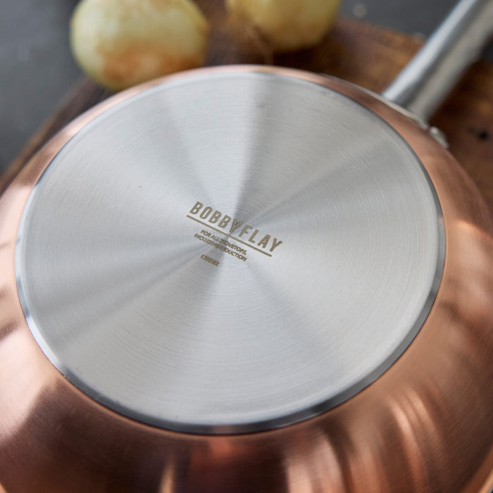 Copper-clad 2-quart chef’s pan with lid on a wooden surface next to onions.