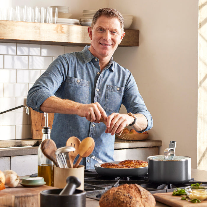Man grinding pepper over food in a GreenPan Bobby Flay 12-inch frypan, with various kitchen items nearby.