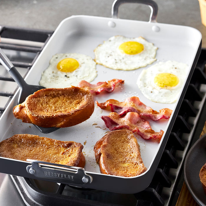 Rectangular nonstick double burner griddle with 3 eggs, 2 bacon strips, and 3 French toast slices cooking.