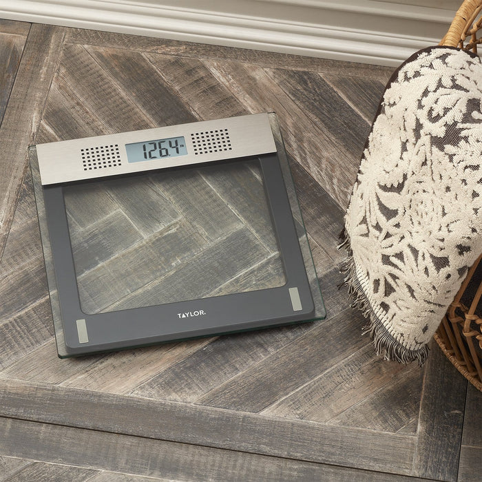 Overhead view of a Taylor digital bathroom scale with a silver display panel on a glass surface, set on a wood floor beside a woven chair.