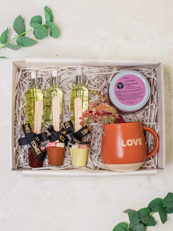 A gift set containing various skincare products and a mug with 'LOVE' written on it, presented in a wooden box lined with shredded paper.