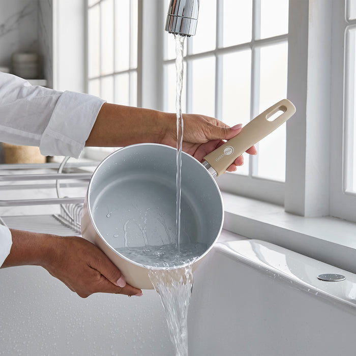 Rinsing a taupe ceramic nonstick saucepan with lid at a kitchen sink near a window.