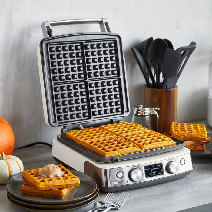 Cream-colored ceramic 4-square waffle maker on a kitchen counter, making Belgian waffles, with plates and pumpkins nearby.