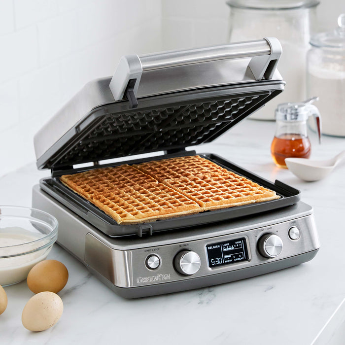 The Elite Ceramic Nonstick 4-Square Waffle Maker | Premiere Stainless Steel sits on a kitchen counter, cooking two golden Belgian waffles amid eggs, batter, flour, and syrup. Its display highlights easy settings and a timer.