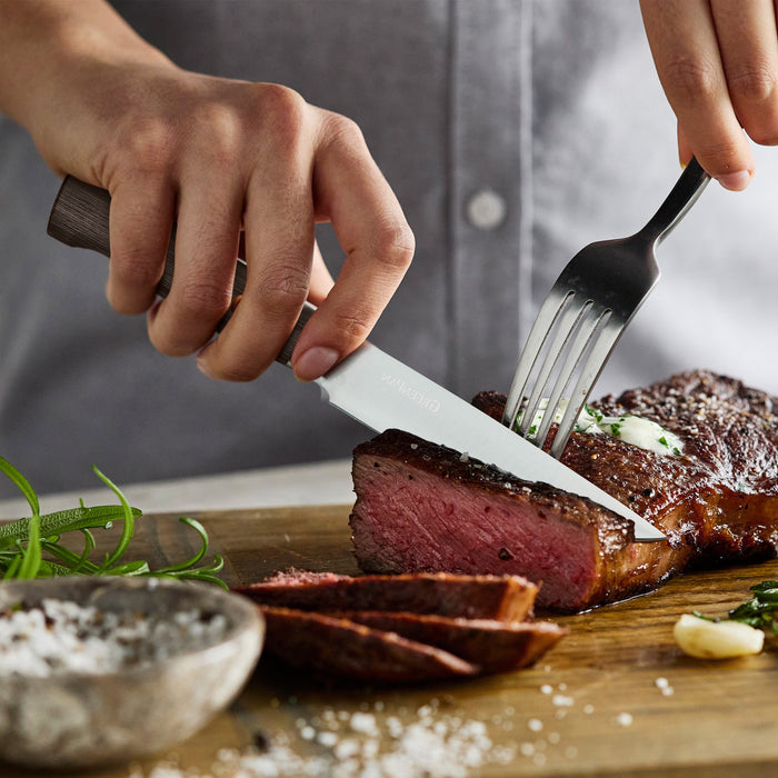 Slicing steak on a wooden board with Elite Cutlery 8-piece steak knife set, dark wood handles, herbs as garnish.