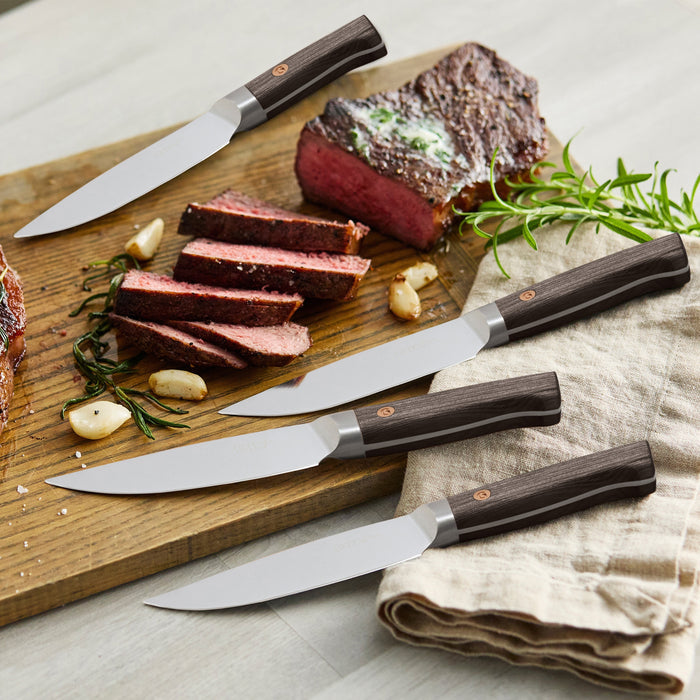 Set of 8 steak knives with dark wood handles on a board, alongside sliced steak and herbs.