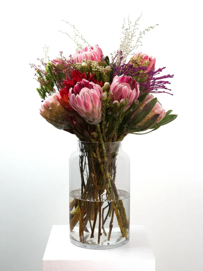 "Bloomable's Girl You Is Fynbos protea bouquet showcased in a modern cylindrical glass vase on a white plinth. Features an eye-catching collection of pink king proteas, vibrant red pincushions, delicate pink filler flowers, and textural greenery with natural woody stems. An exquisite South African fynbos arrangement ideal for Women's Day celebrations or stunning home décor with same-day delivery across South Africa."