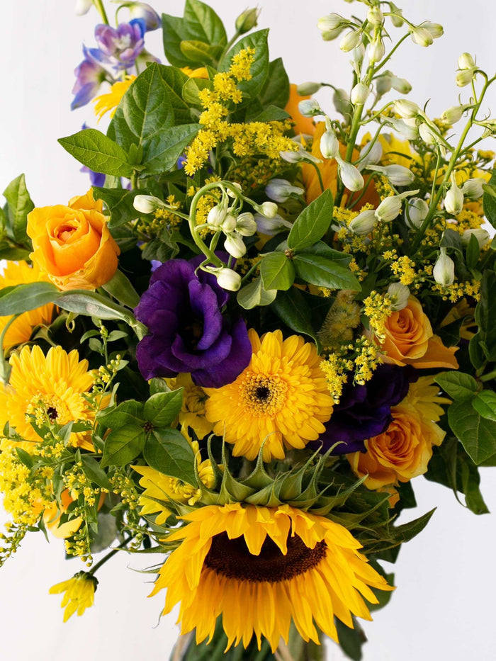 Colorful bunch of seasonal flowers in blues and yellows with sunflowers, roses, and foliage in a vibrant arrangement.