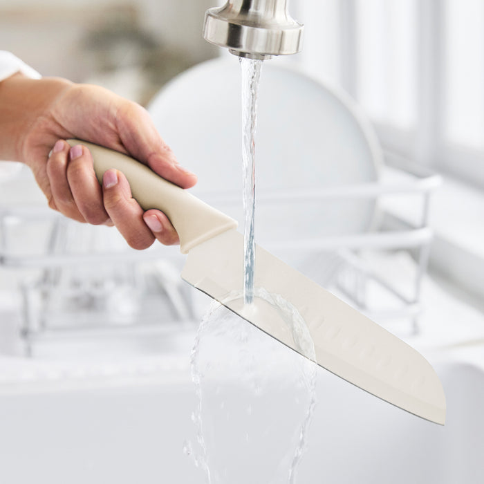 Hand rinsing taupe GreenLife kitchen knife under running sink water, other dishes visible in the background.