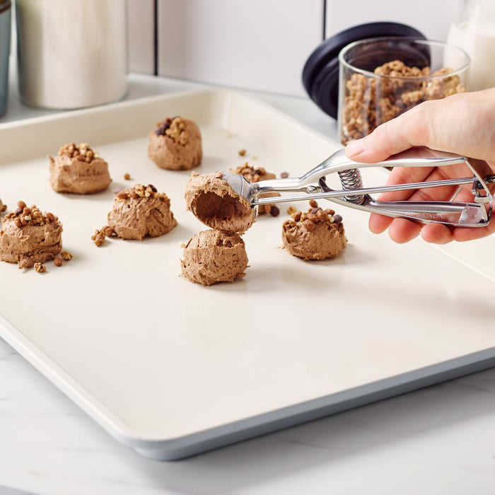 Hand scooping chocolate cookie dough onto a GreenLife ceramic nonstick baking sheet, topping with chocolate bits.