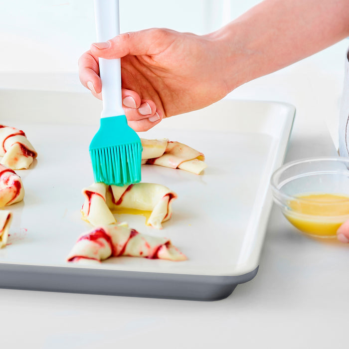 Silicone brush spreading egg wash on pastry dough placed on a gray ceramic nonstick cookie sheet.