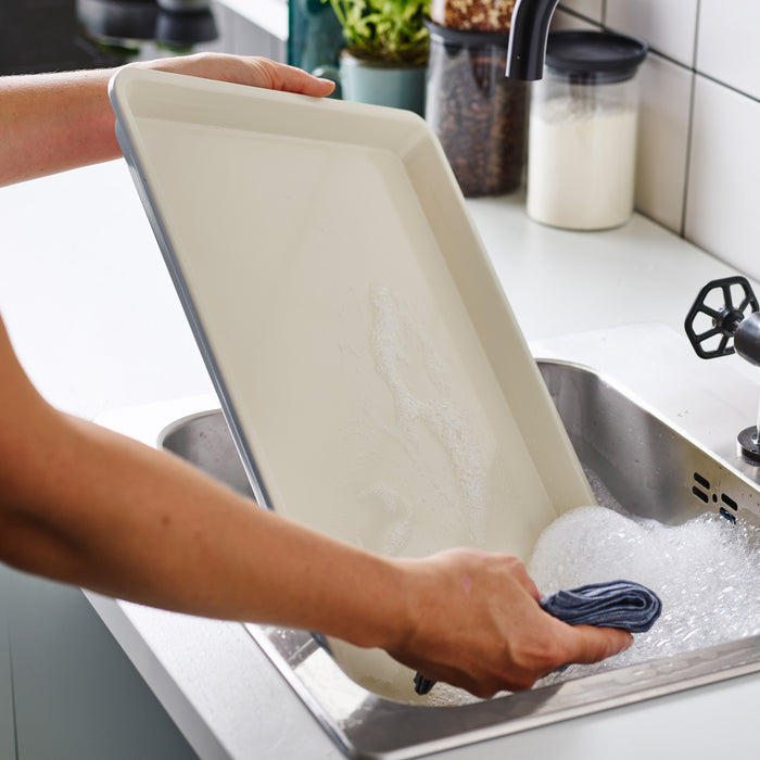 Washing a quartz gray GreenLife ceramic nonstick cookie sheet in a soapy sink with a cloth.