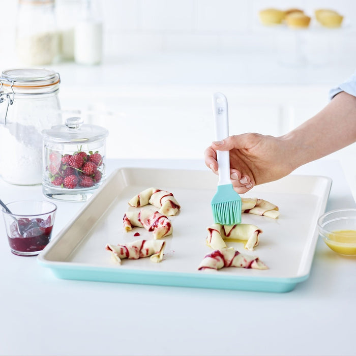 Hand buttering pastry rolls on a turquoise ceramic nonstick cookie sheet; jam and berries on the side.