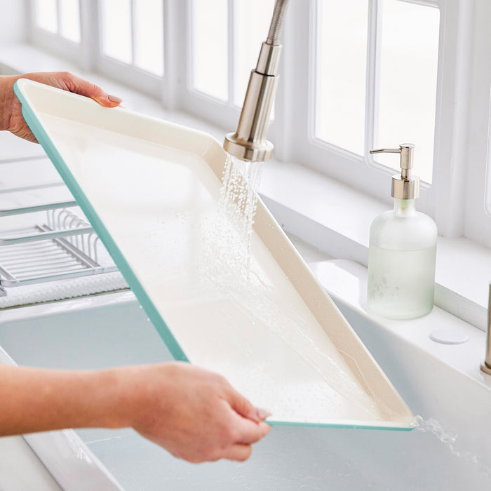 Person washing a turquoise sheet pan under running water in a kitchen sink.