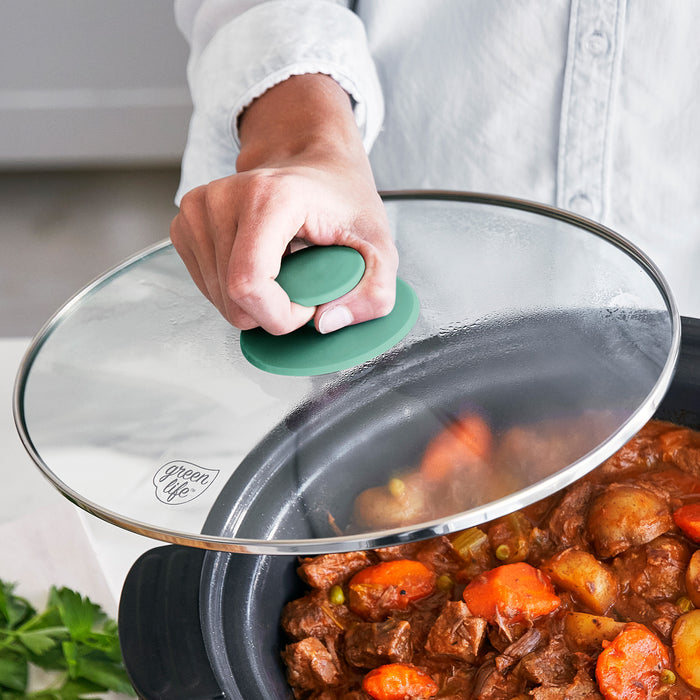 Glass lid being placed on a pot of stew with visible carrots and beef, held by a person.