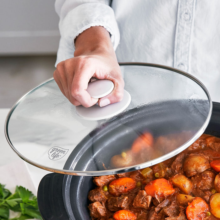 Person holding a glass lid on a slow cooker with stew inside, featuring carrots and other vegetables.