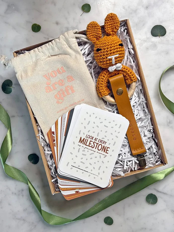 A gift box set on a white marble surface, featuring a cream drawstring pouch with pink text, a crocheted orange bunny keychain with a brown leather loop, a stack of white cards, and shredded paper inside a kraft box, with green ribbon and eucalyptus leaves around it.
