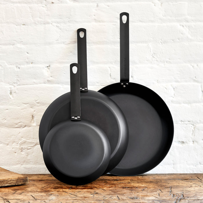 Three carbon steel frying pans in different sizes stacked upright on a wooden surface against a white brick wall.