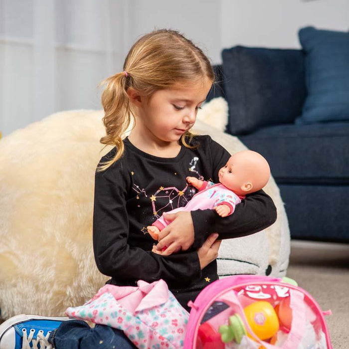 A young girl with blonde pigtails wears a black constellation shirt, sitting indoors and holding the My Sweet Baby Dlx Baby Ensemble by My Sweet Baby. Toys and a pink bag are in front, making this set perfect for hours of pretend play.