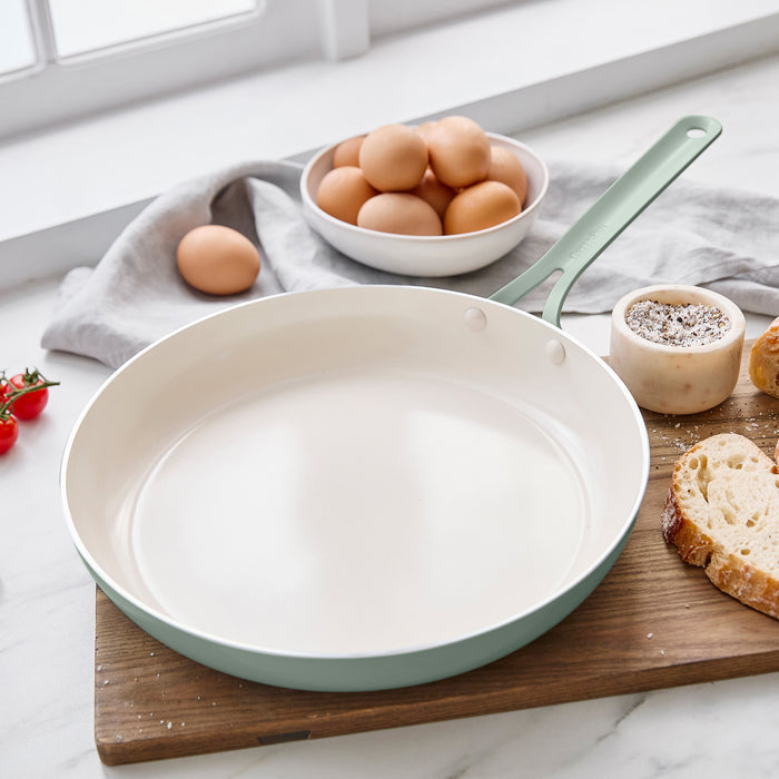 Light green frying pan on a wooden cutting board with eggs and bread in the background.
