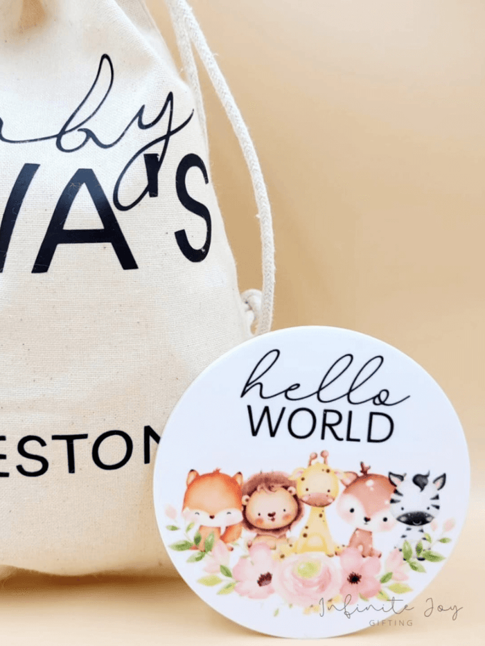 Personalised Baby Milestone Disc with "Hello World" message and animal design beside a cotton drawstring bag.